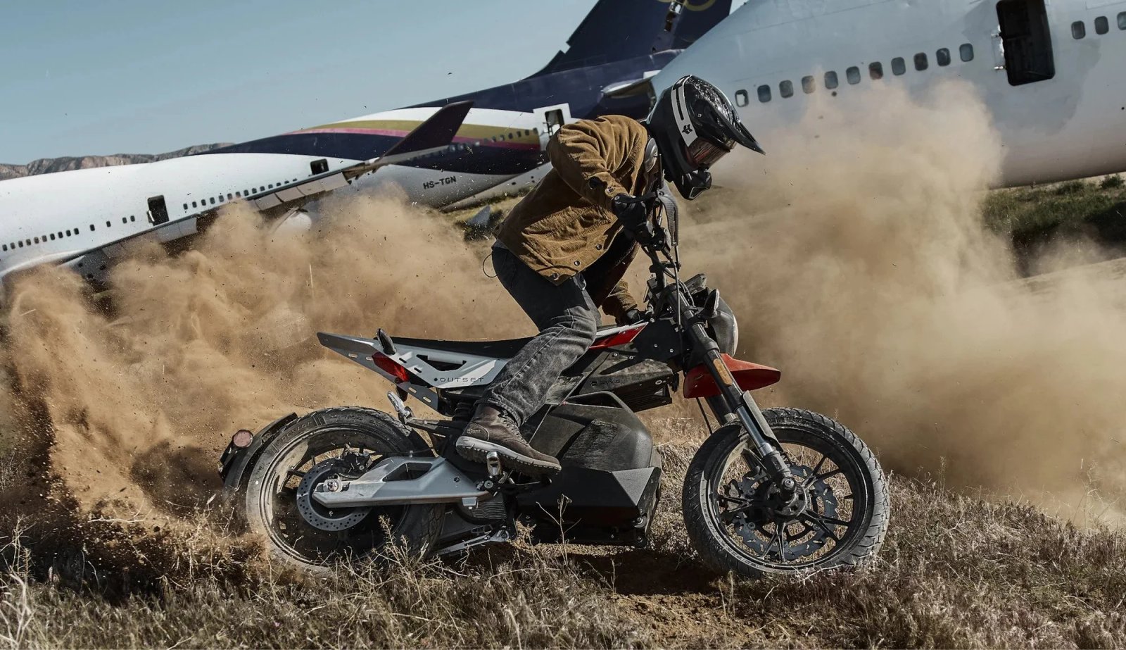 Persona conduciendo una motocicleta en un terreno polvoriento con un avión abandonado al fondo.